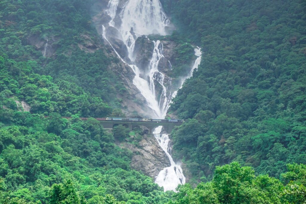 Scenic view of the impressive Dudhsagar Waterfall, surrounded by lush greenery, with a prominent railway bridge in the foreground.