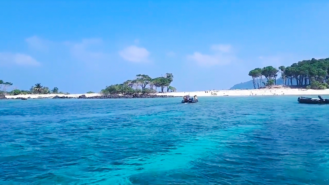 The boat approaches the shore of North Cinque Island, where the stunning colors of the water blend the deep navy blue of the sea with the turquoise hues of the lagoon, all against the backdrop of the island's ultra-white sandy shore and the lush littoral forests.