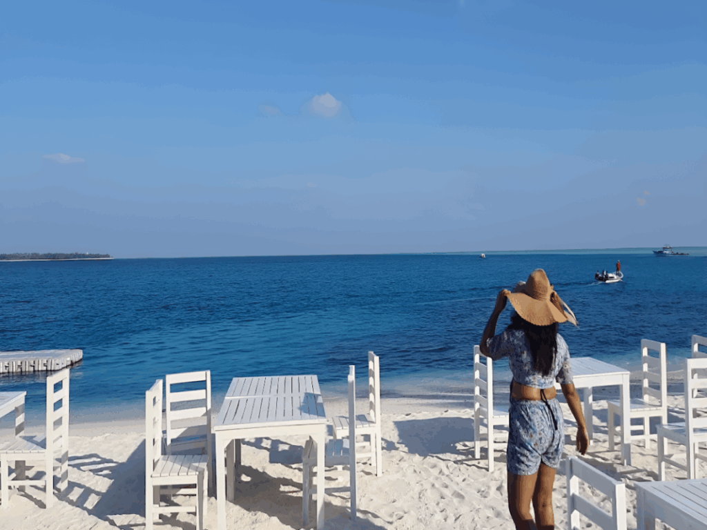 6 Nights 7 Days Agatti-Kavaratti-Bangaram-Thinnakara Affordable Luxury Lakshadweep Package by SetMyTrip – White sandy beach with oceanfront dining setup and tourist enjoying the sea view.